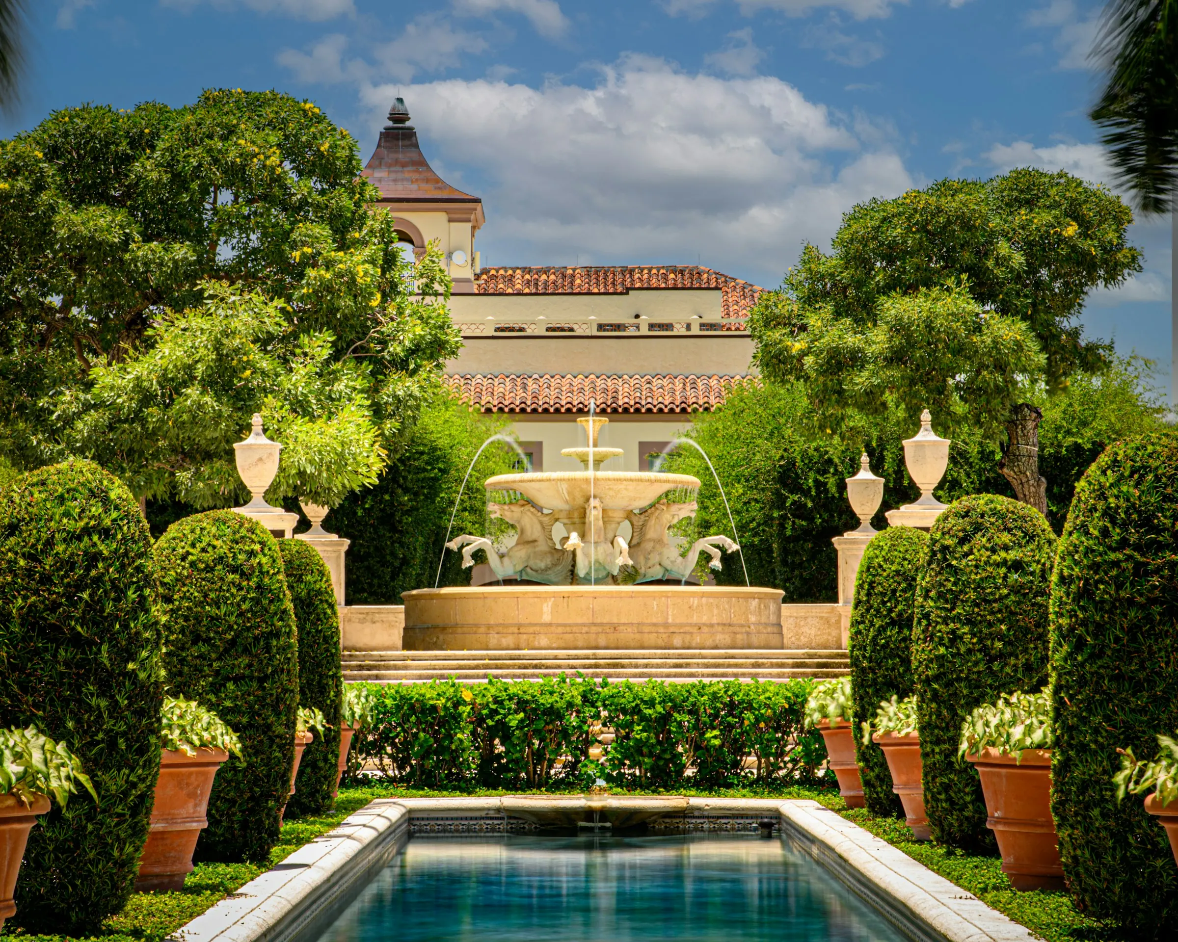 A formal garden with a fountain and clipped hedges before a Palm Beach hotel